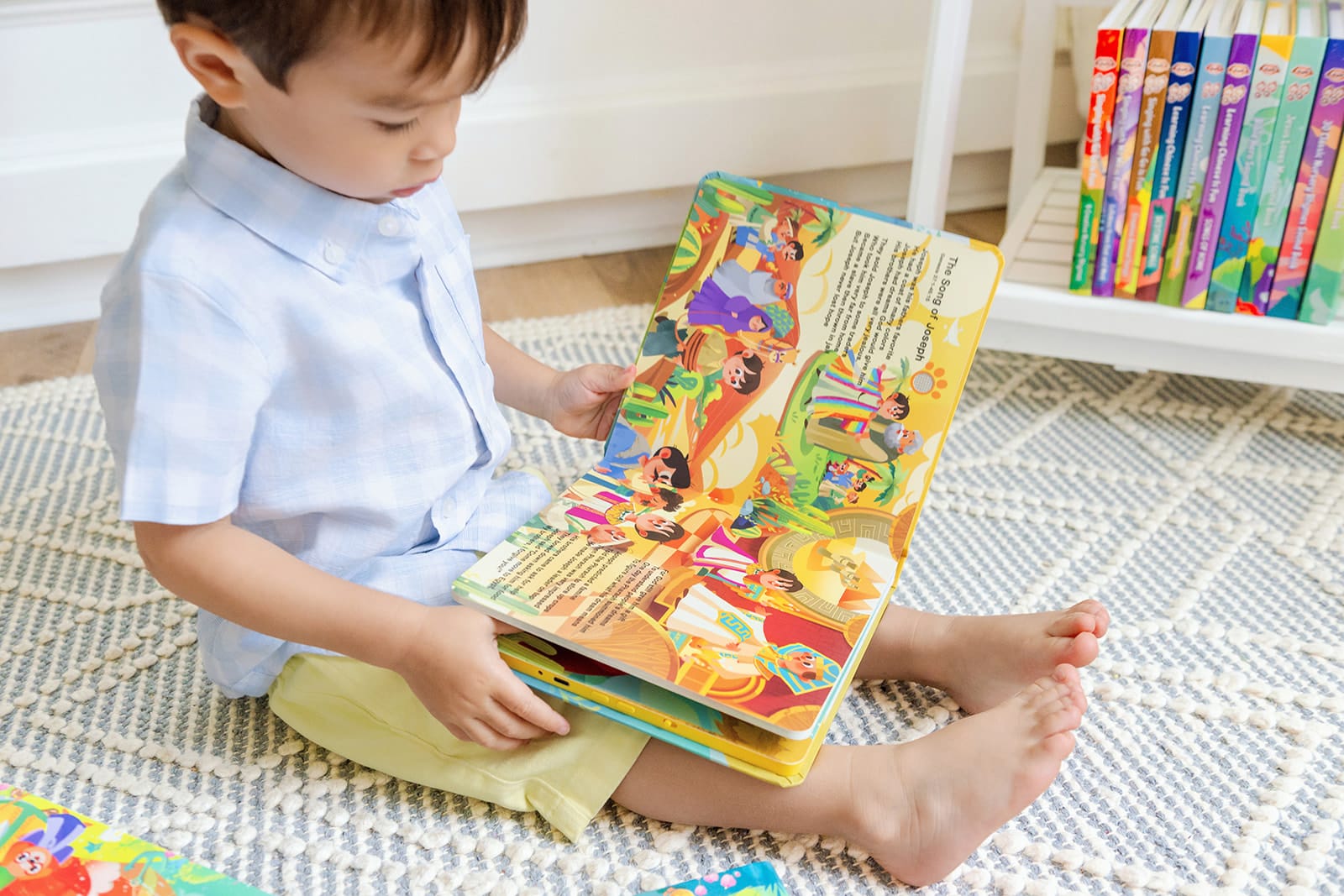 A toddler boy sitting on a rug reading a Go Go Bible Hero Stories board book entirely on his own, open to the illustrated 'The Song of Joseph' page, with a shelf of Go Go Christian sound books behind him — no screen involved